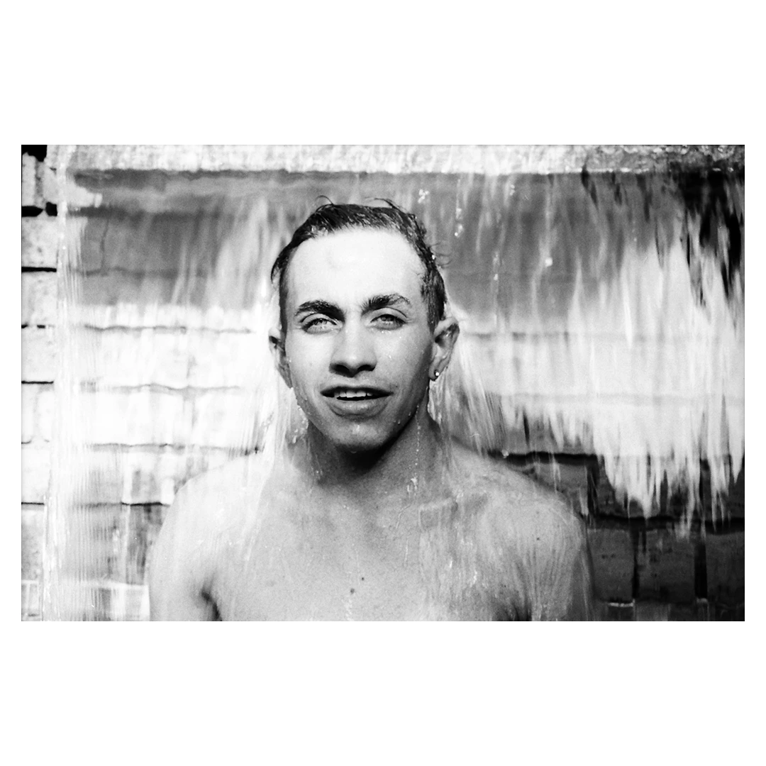 Black and white portrait of a smiling young man standing shirtless under a sheet of falling water against a brick wall